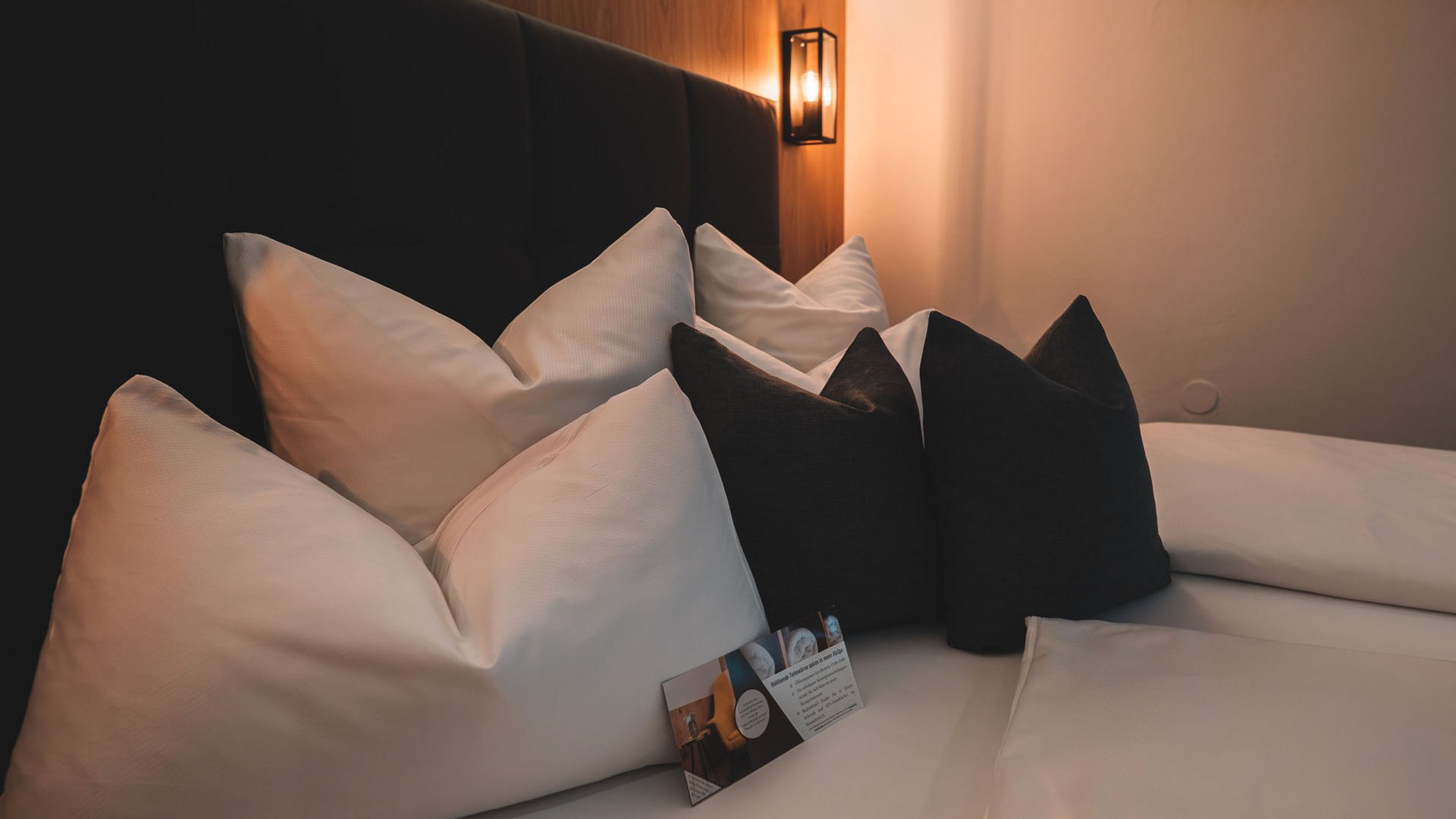 4-Sterne-Hotel Stubaital: Ihr Alphof erwartet Sie! Gemütliches Bett mit weißen und dunklen Kissen und warmem Wandlicht