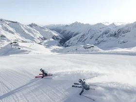 Two skiers descend through deep snow in front of snow-covered mountain peaks
