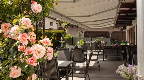 Einblicke in unser Hotel im Stubaital in Fulpmes Sonnige Terrasse mit Tischen und Stühlen unter Markisen neben blühendem Rosenbogen