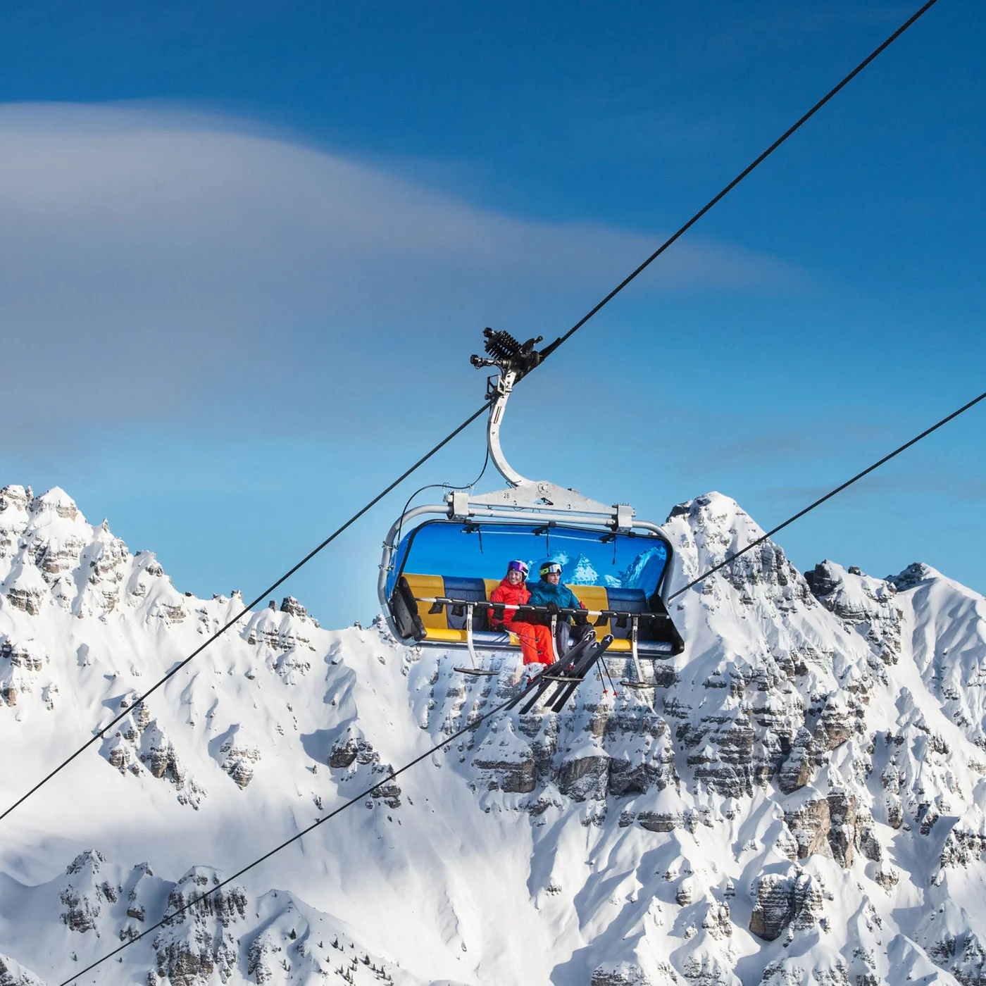 Open chairlift carrying two skiers above snow-covered mountain peaks