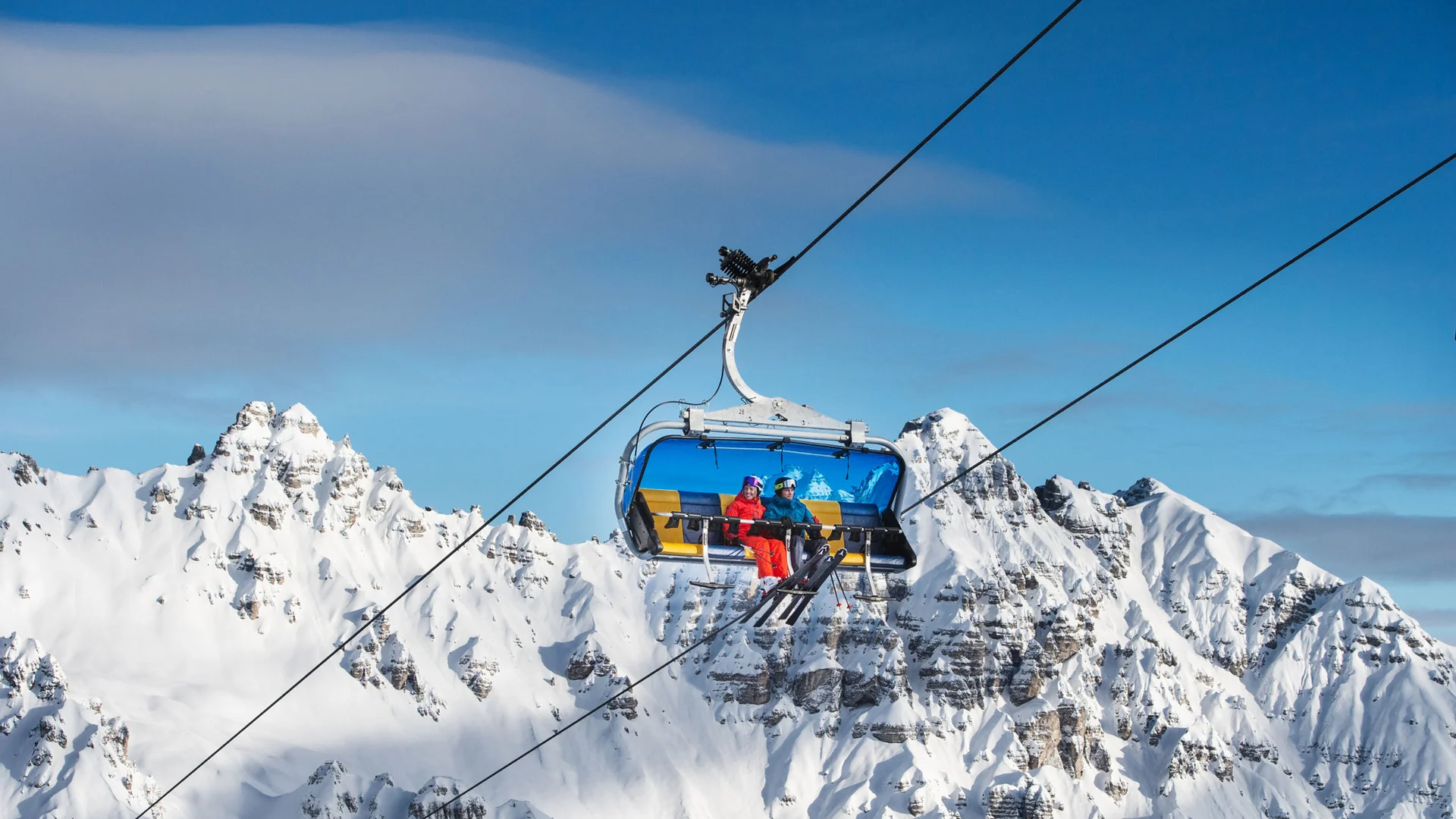 Open chairlift carrying two skiers above snow-covered mountain peaks