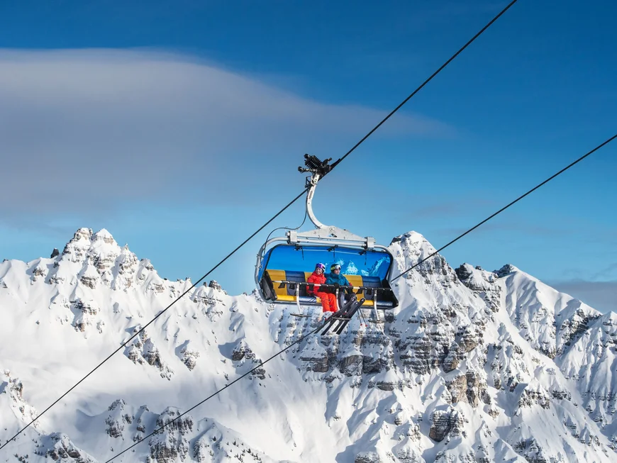 Open chairlift carrying two skiers above snow-covered mountain peaks