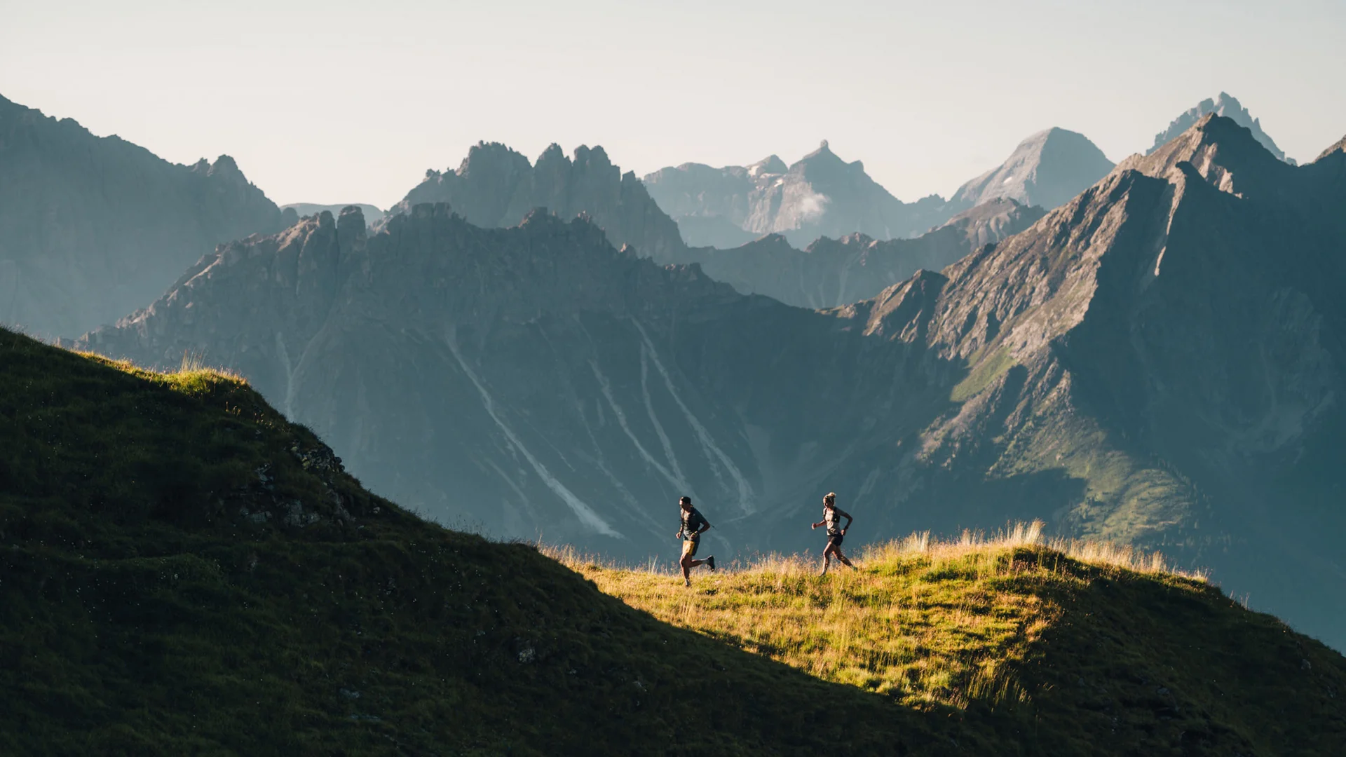 Two runners on a sunlit mountain ridge with a dramatic mountain landscape