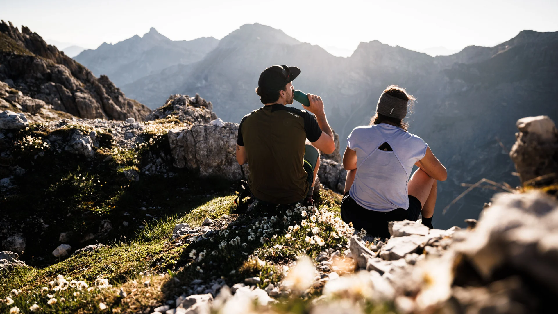 Two hikers sit on a mountain ridge overlooking the Alps