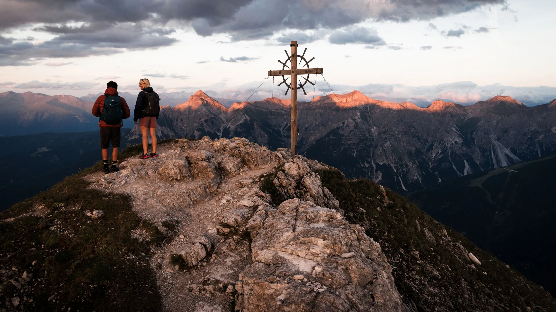 Two hikers and a summit cross overlooking mountains in evening light
