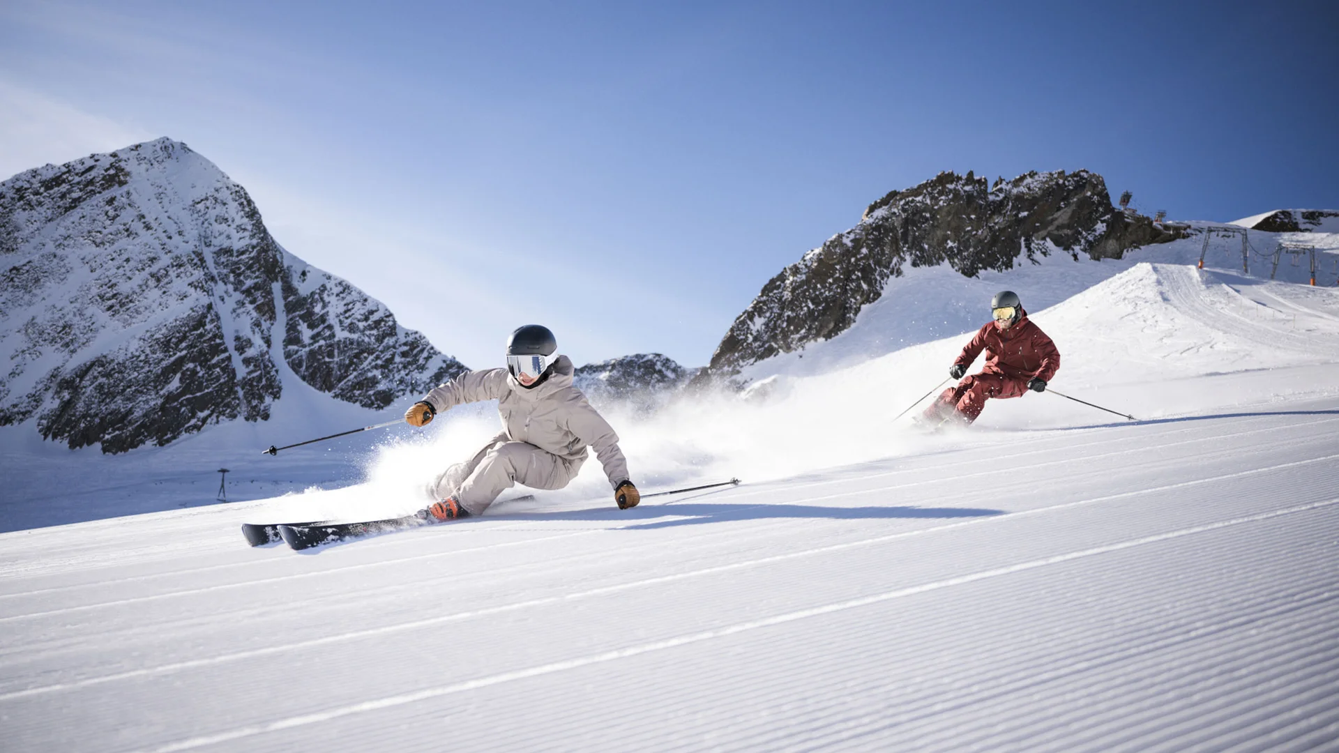 Two skiers descend a snowy slope in front of rocky mountains