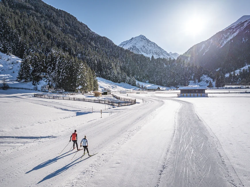 Two cross-country skiers on a snowy trail in a mountain valley under sunny skies