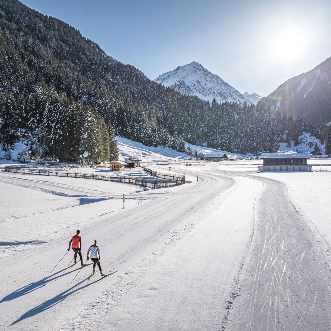 Two cross-country skiers on a snowy trail in a mountain valley under sunny skies