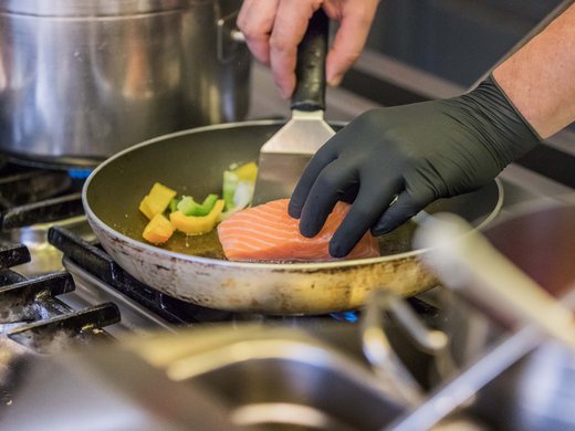 Hotel Alphof: ein Gourmethotel in Tirol Hand mit schwarzem Handschuh brät Lachs und Gemüse in einer Pfanne auf dem Herd