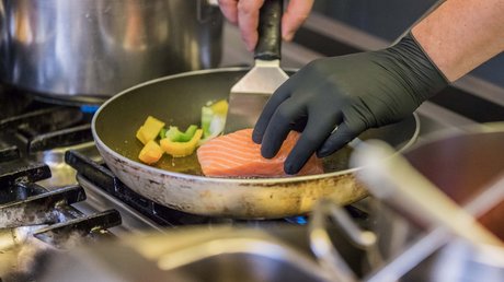 Einblicke in unser Hotel im Stubaital in Fulpmes Hand mit schwarzem Handschuh brät Lachs und Gemüse in einer Pfanne auf dem Herd