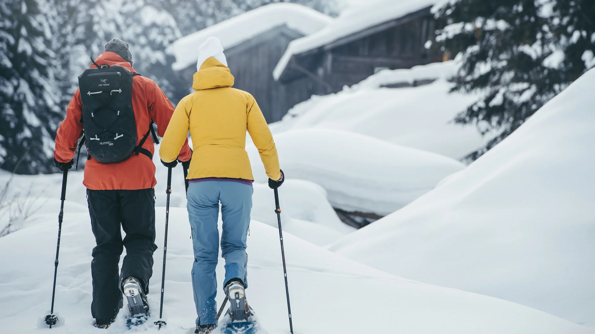 Two cross-country skiers trek through snowy scenery toward a mountain hut