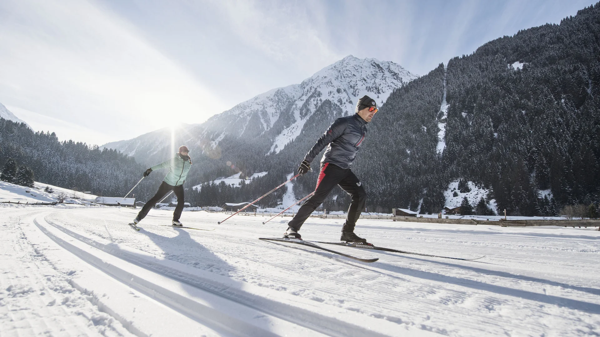 Two cross-country skiers on a snowy trail with mountains in the background