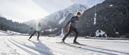 Two cross-country skiers on a snowy trail with mountains in the background