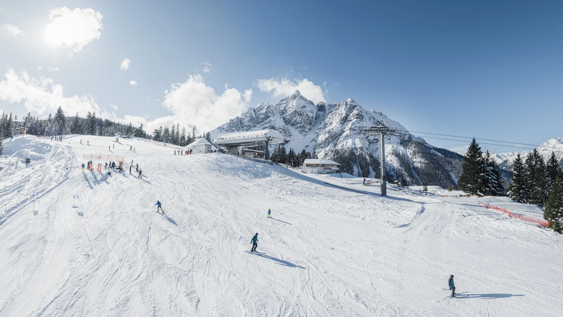 Skiers on a snowy slope with a cable car and snow-covered mountains