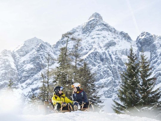 Winterurlaub in Tirol: Hotel Alphof in Fulpmes Zwei Menschen auf Schlitten vor schneebedeckten Bergen und Tannenbäumen