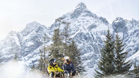 Einblicke in unser Hotel im Stubaital in Fulpmes Zwei Menschen auf Schlitten vor schneebedeckten Bergen und Tannenbäumen
