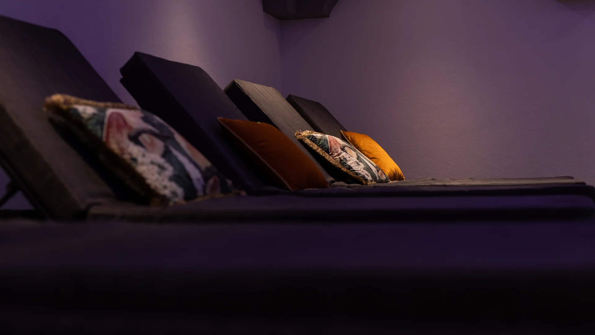 Dark seating or lounging area with colorful cushions against a purple-lit wall