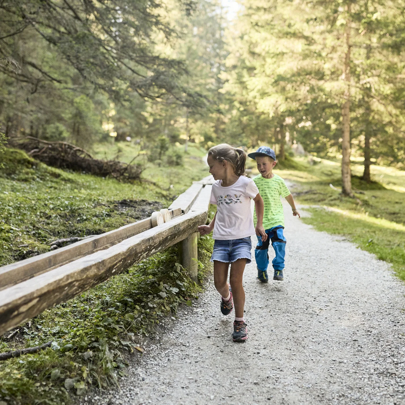Two children walk along a forest path beside a wooden railing