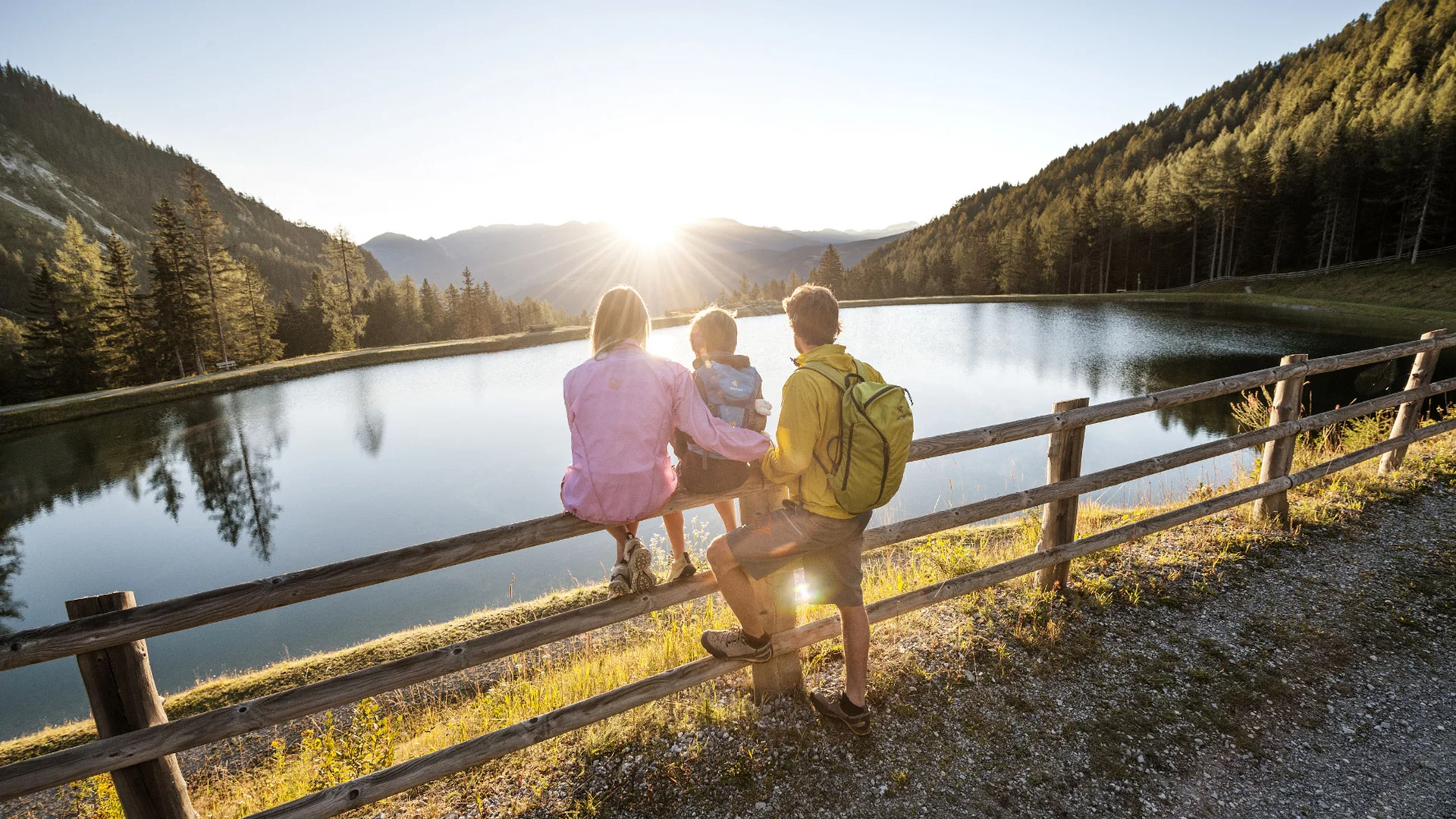 Family sitting on a wooden fence beside a mountain lake at sunset