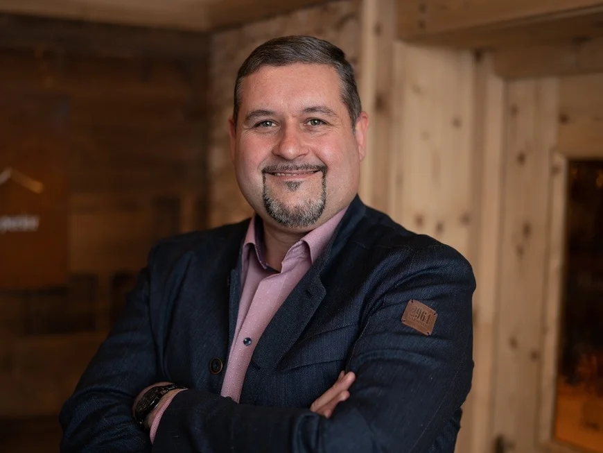 Portrait of a smiling man in a dark suit inside a rustic wooden cabin