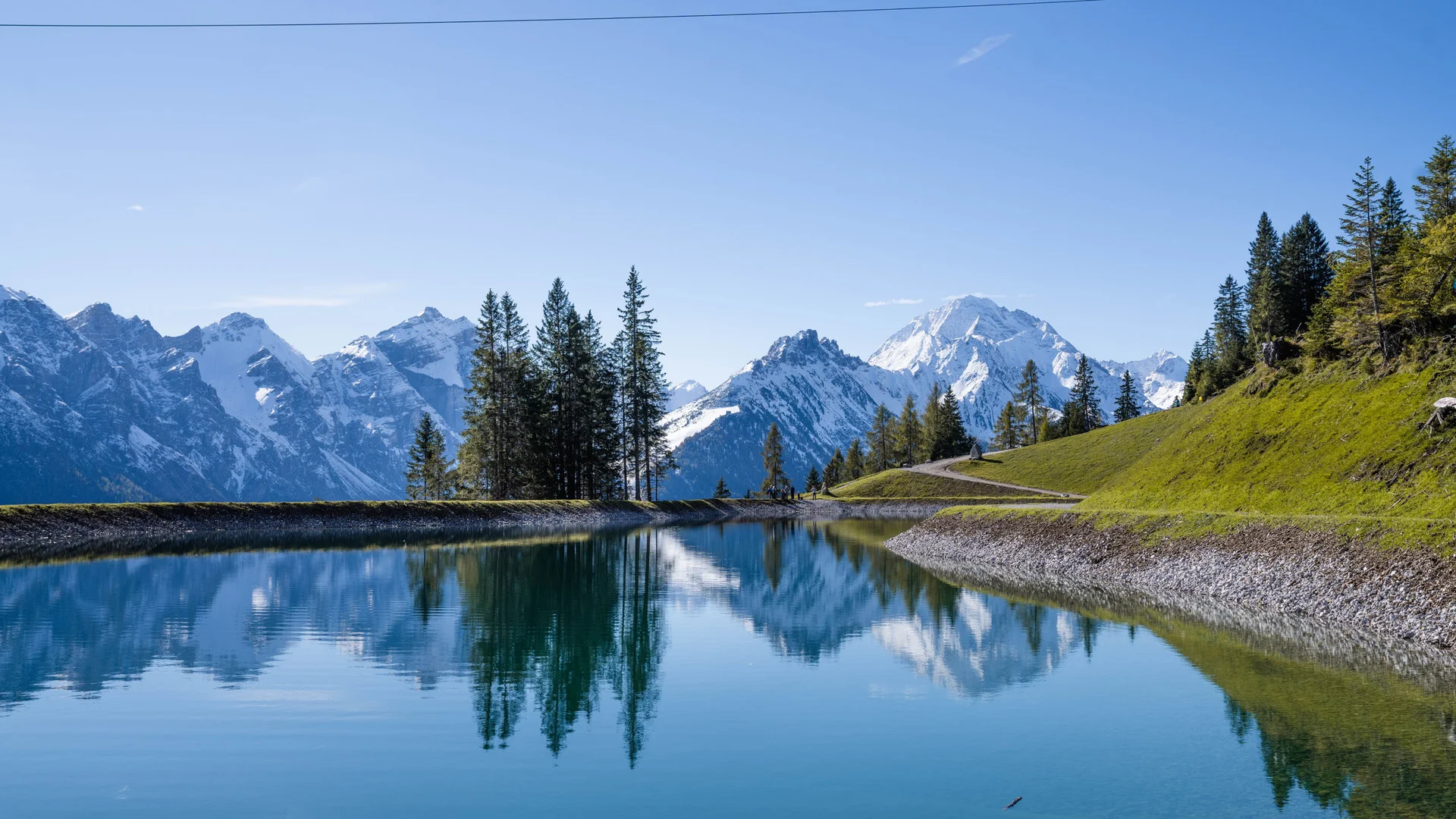 Mountain lake reflecting conifer trees and snow-capped Alps under a blue sky