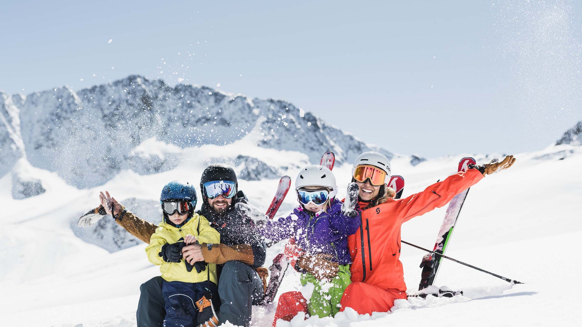 Familie mit Skihelm und Skiausrüstung sitzt lachend im Schnee vor Bergkulisse