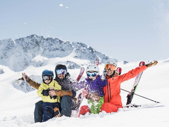 Winterurlaub in Tirol: Hotel Alphof in Fulpmes Familie mit Skihelm und Skiausrüstung sitzt lachend im Schnee vor Bergkulisse