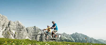Two cyclists on a flower-filled mountain meadow with rugged peaks behind