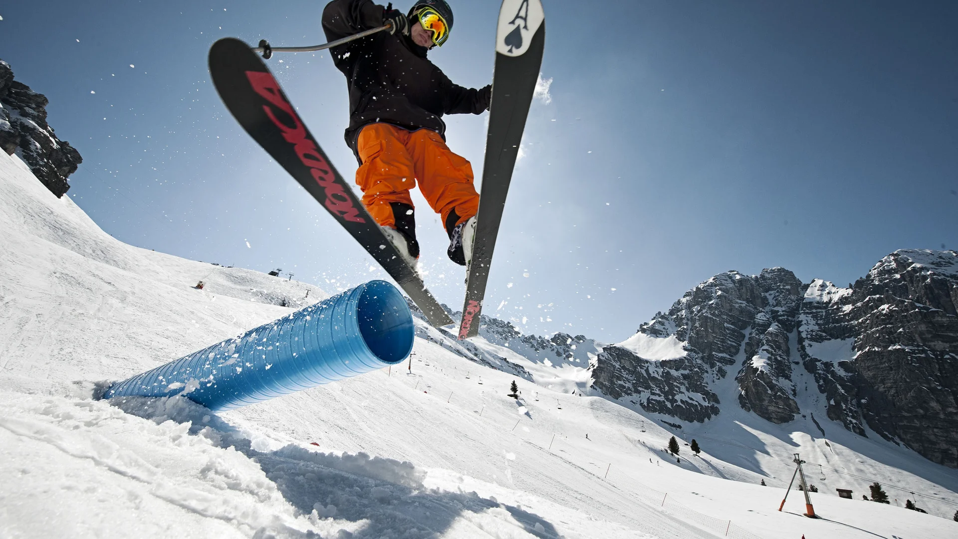 Skier jumping over a blue snow tube in front of snowy mountain peaks