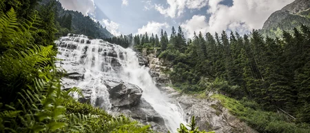 Wide waterfall in a forested mountain valley beneath a cloudy sky