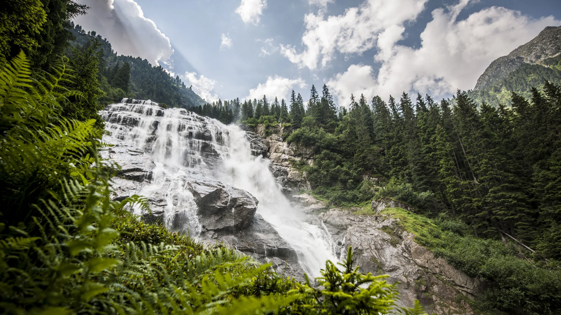 Wide waterfall in a forested mountain valley beneath a cloudy sky