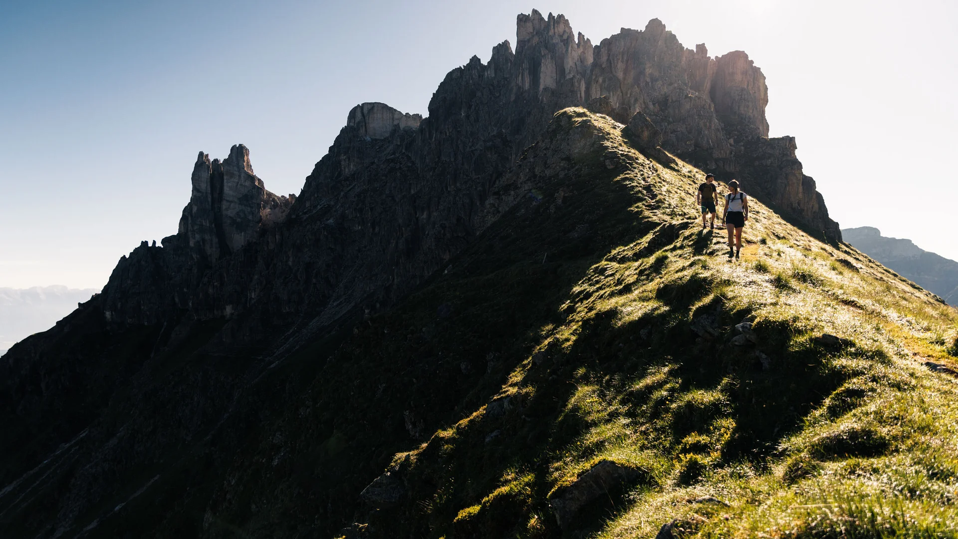 Two hikers on a sunlit mountain ridge with a rugged rocky backdrop