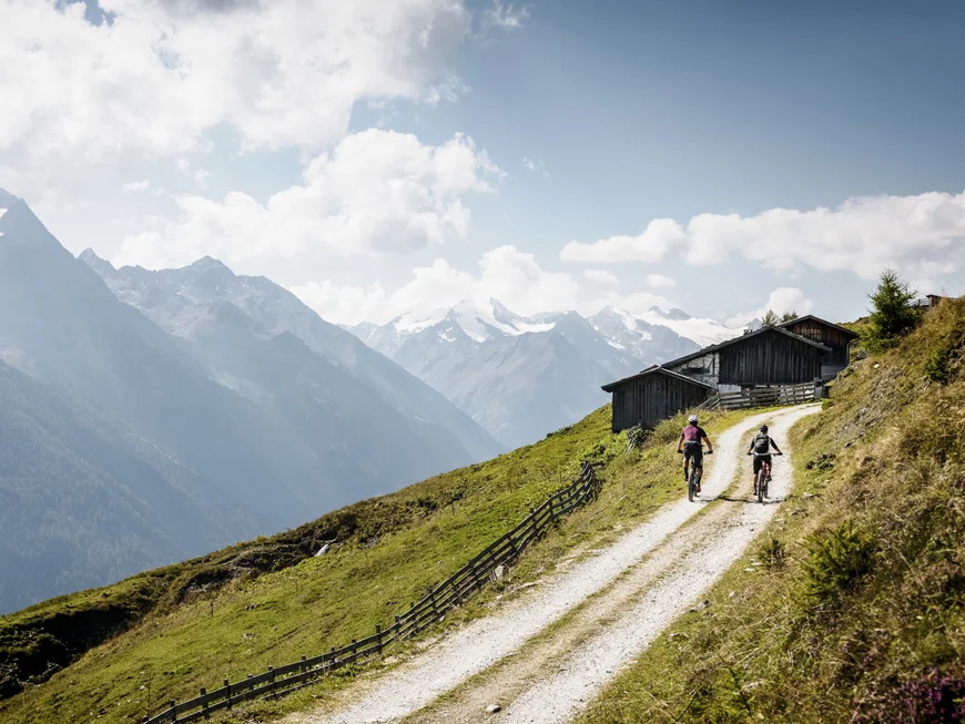 Two cyclists on a mountain trail with snowy Alps and mountain huts