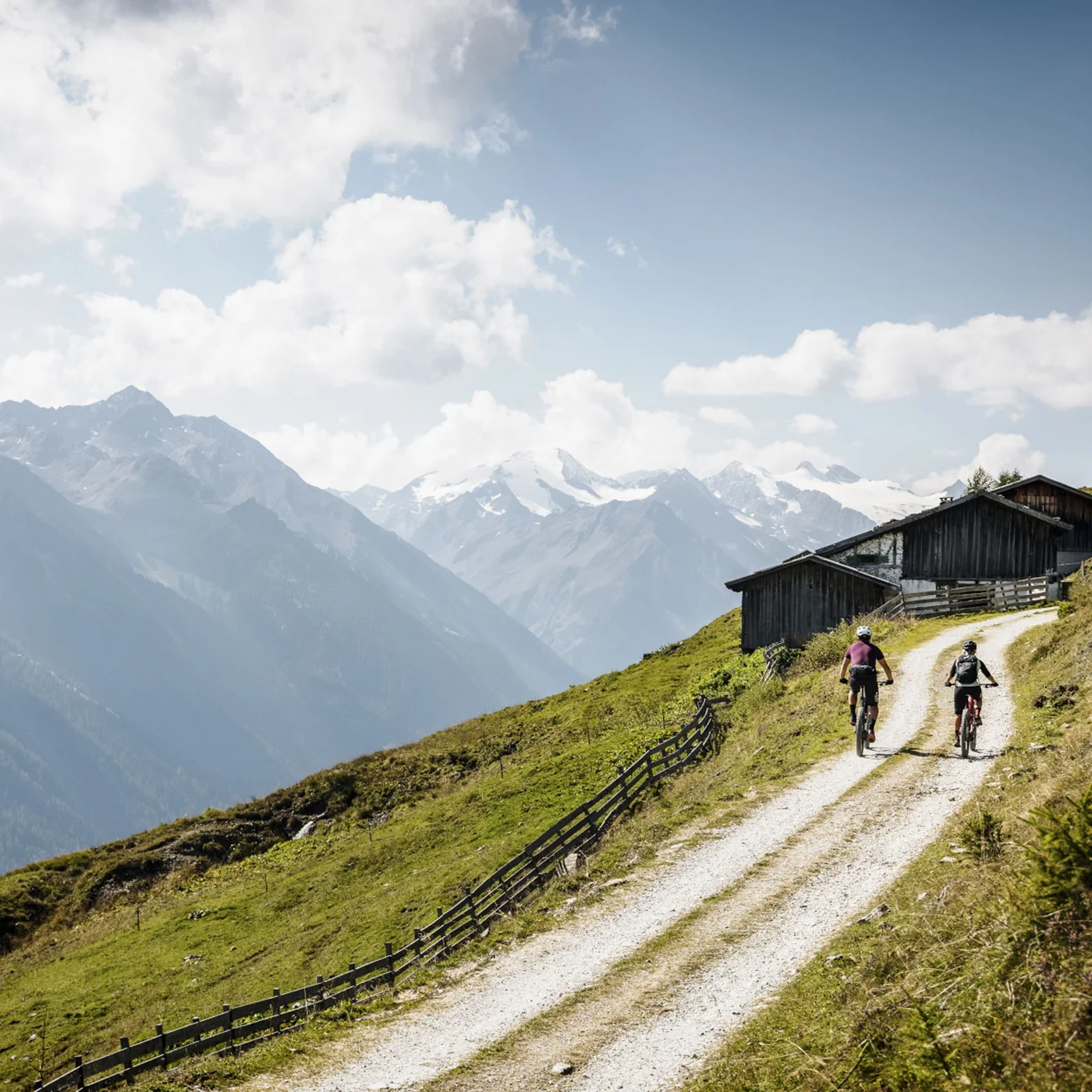 Two cyclists on a mountain trail with snowy Alps and mountain huts
