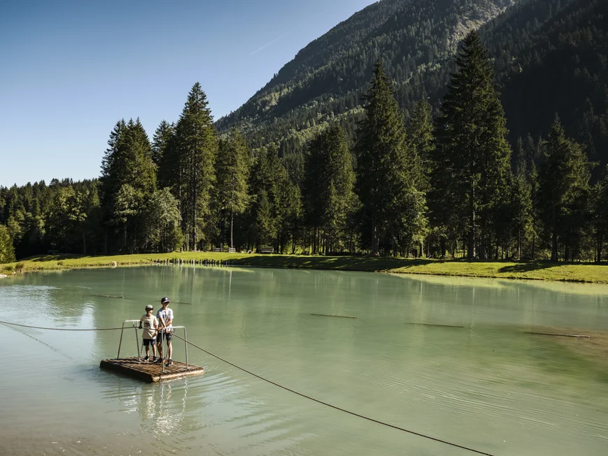 Two people on a raft ferry crossing a lake beneath a forested mountain