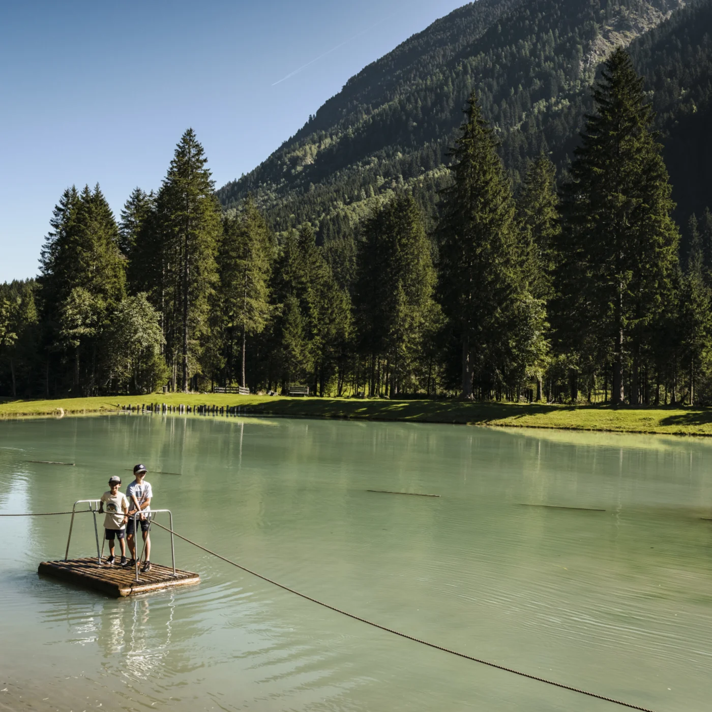 Two people on a raft ferry crossing a lake beneath a forested mountain