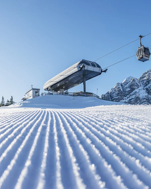 Groomed ski slope with chairlift and cable car gondola before snow-covered mountain peaks