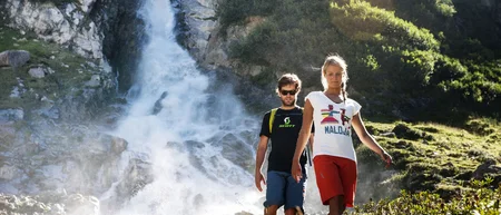 Two hikers walking in front of a rushing waterfall in a rocky mountain landscape