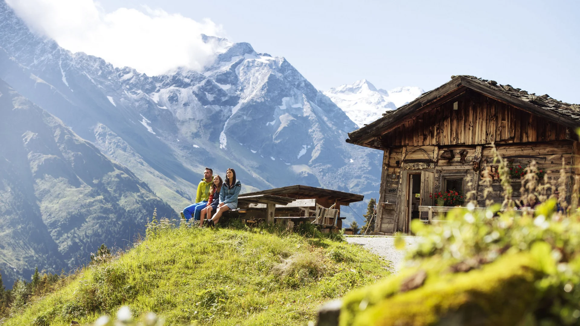 Three people outside a rustic mountain hut with snow-covered Alps in the background