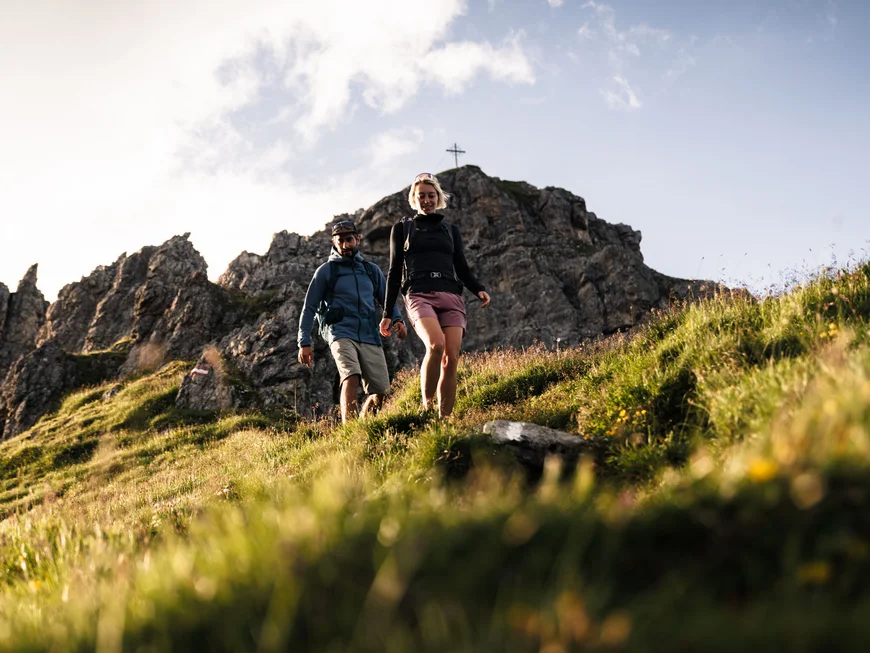 Hotel Alphof | your basecamp for walking holidays in Austria Two hikers walk across a meadow in front of a rocky mountain landscape