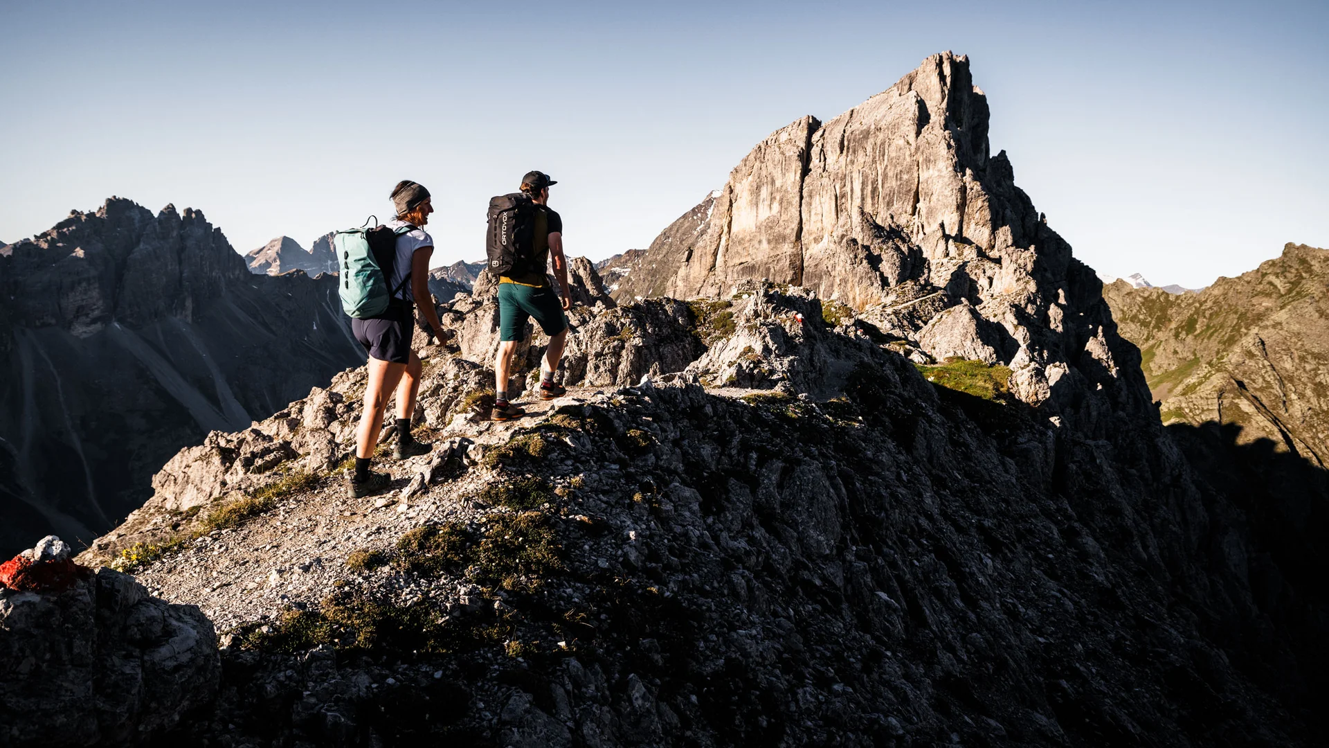 Two hikers on a rocky mountain ridge below a distinctive peak