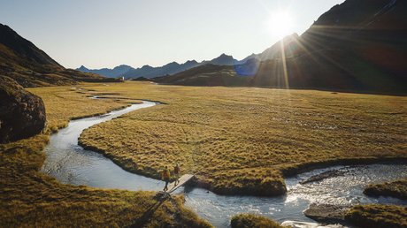 Einblicke in unser Hotel im Stubaital in Fulpmes Zwei Wanderer überqueren bei Sonnenuntergang einen kleinen Holzsteg in einer Bergwiese.
