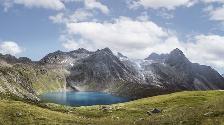 Einblicke in unser Hotel im Stubaital in Fulpmes Bergsee umgeben von grünen Wiesen und steinigen Bergen unter bewölktem Himmel