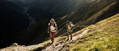Two hikers on a mountain trail overlooking an alpine valley and snow-capped peaks