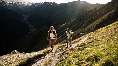 Two hikers on a mountain trail overlooking an alpine valley and snow-capped peaks