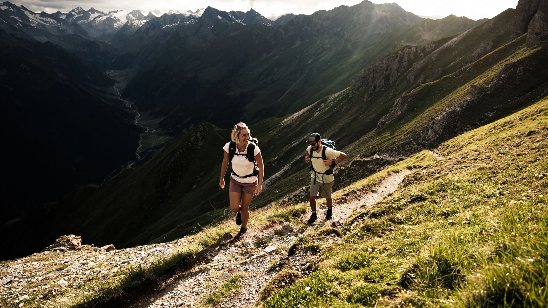 Two hikers on a mountain trail overlooking an alpine valley and snow-capped peaks