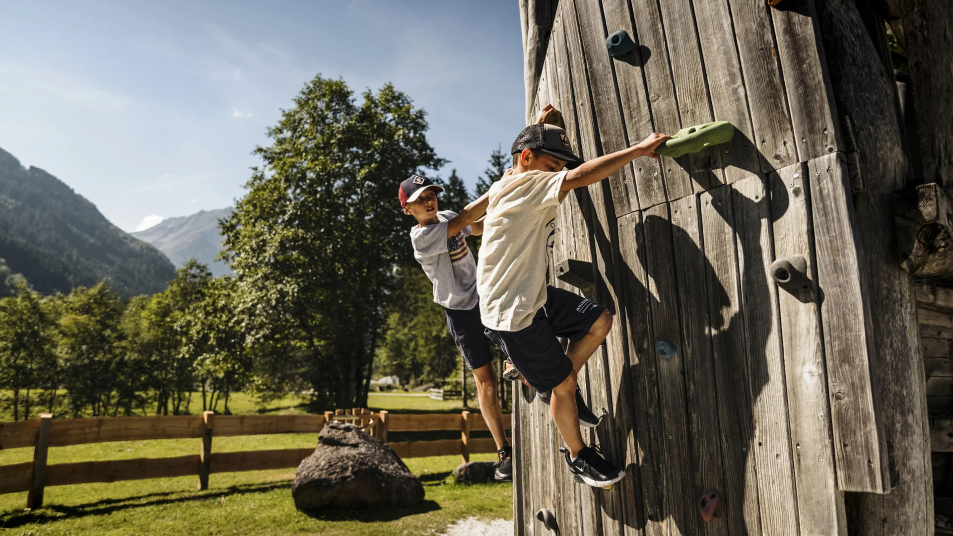 Two children climb a wooden climbing wall against a mountain backdrop