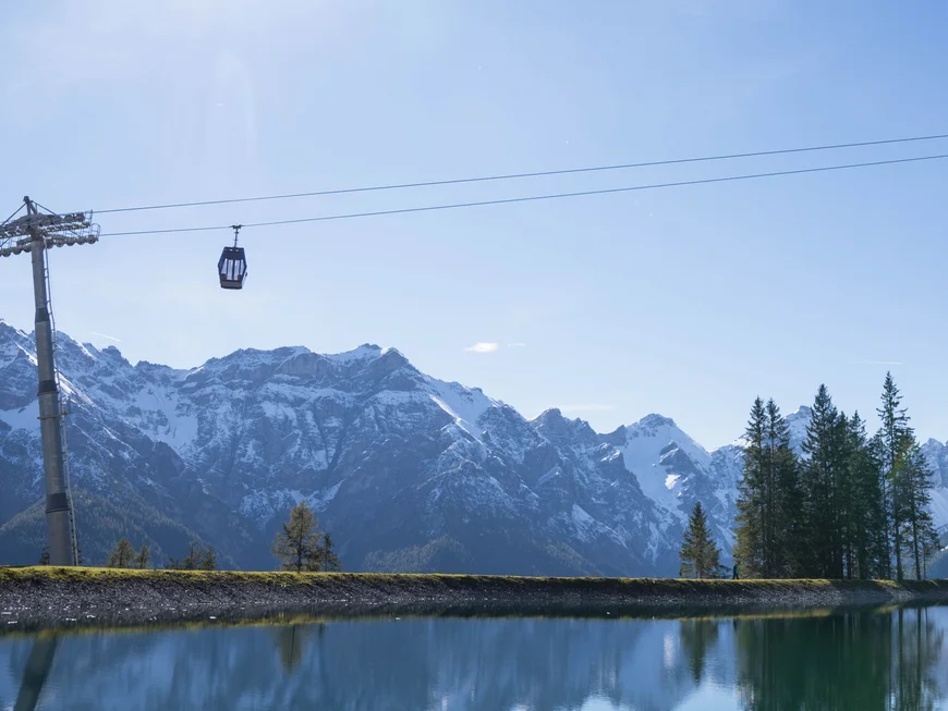 Mountain lake reflecting snowy Alps, with a cable car and pine trees