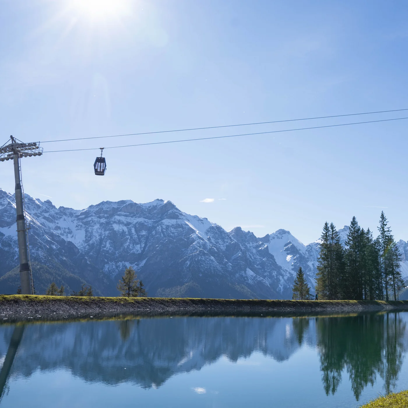 Mountain lake reflecting snowy Alps, with a cable car and pine trees
