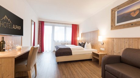 Einblicke in unser Hotel im Stubaital in Fulpmes Modernes Hotelzimmer mit Doppelbett, Schreibtisch, Sofa und Balkon mit roten Vorhängen